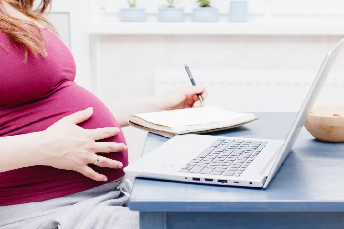 Mloda kobieta w ciazy uczy sie przy laptopie i robi notatki w zeszycie