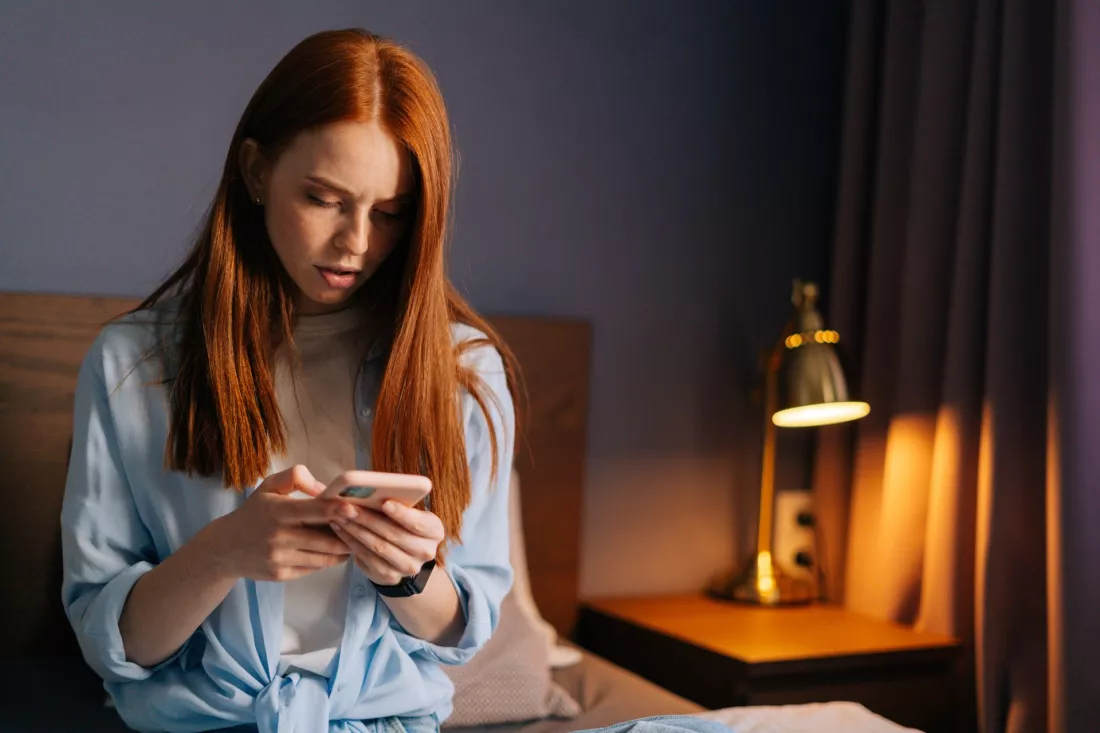 Młoda kobieta z poważną miną pisze wiadomość w telefonie w swoim pokoju