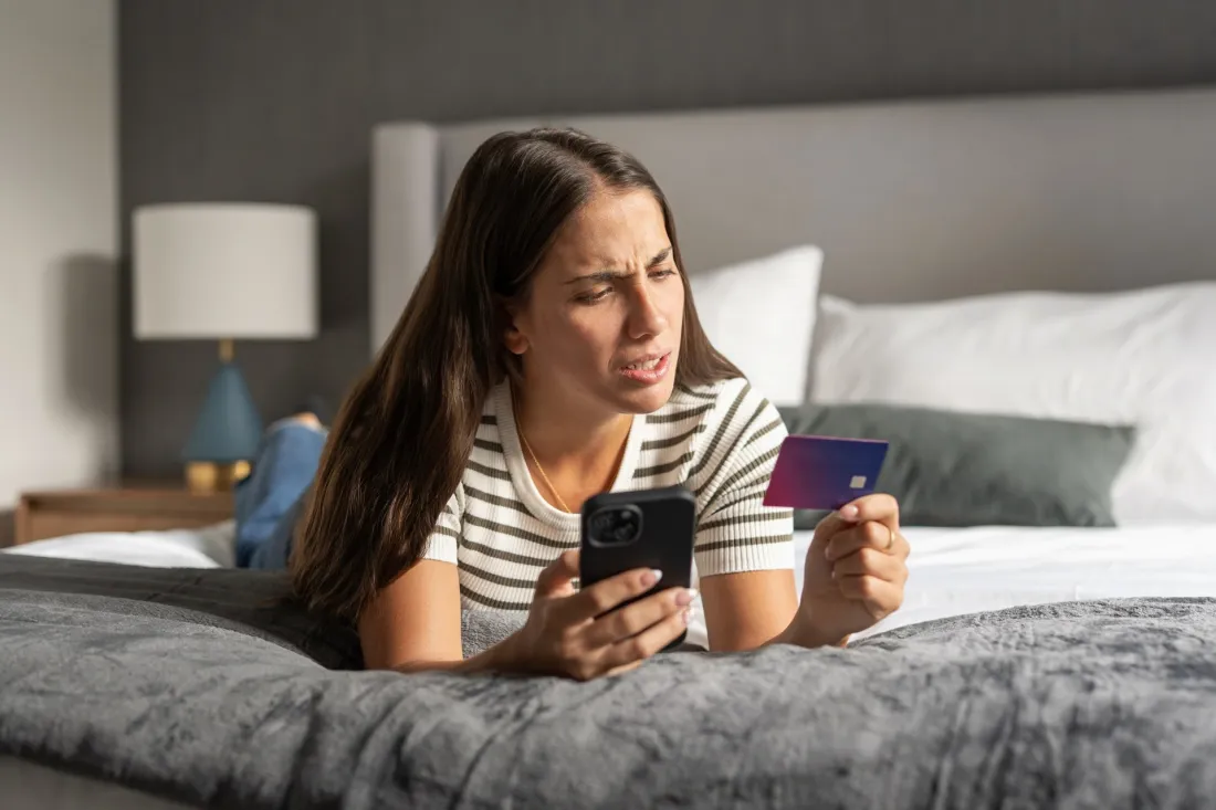 Zaniepokojona młoda kobieta leży na łóżku i patrzy na smartfon, w drugiej ręce trzyma kartę płatniczą.